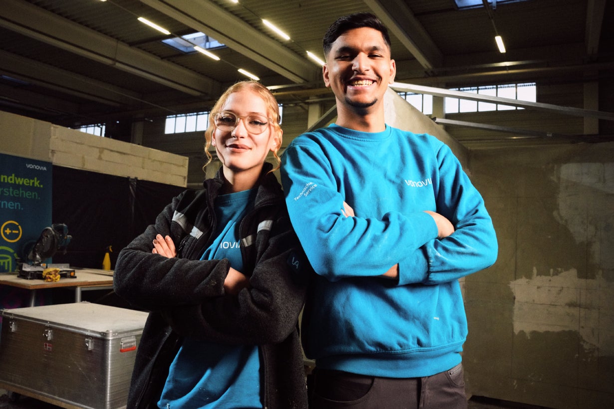 2 Auszubildende in Arbeitskleidung stehen mit verschränkten Armen und lächeln.