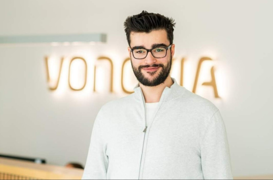 Mann steht lächelnd in Büro vor beleuchtetem Vonovia-Zeichen.