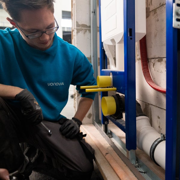 Ein Auszubildender mit einem Schraubschlüssel in der Hand kniet vor einer Anlage.
