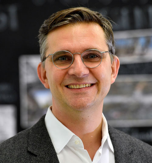 Mann lächelt in Kamera, trägt Brille, Anzug und Hemd, Innenraum.