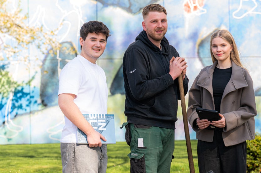Drei Auszubildende aus dem kaufmännischen und handwerklichen Bereich stehen in einer Grünanlage.
