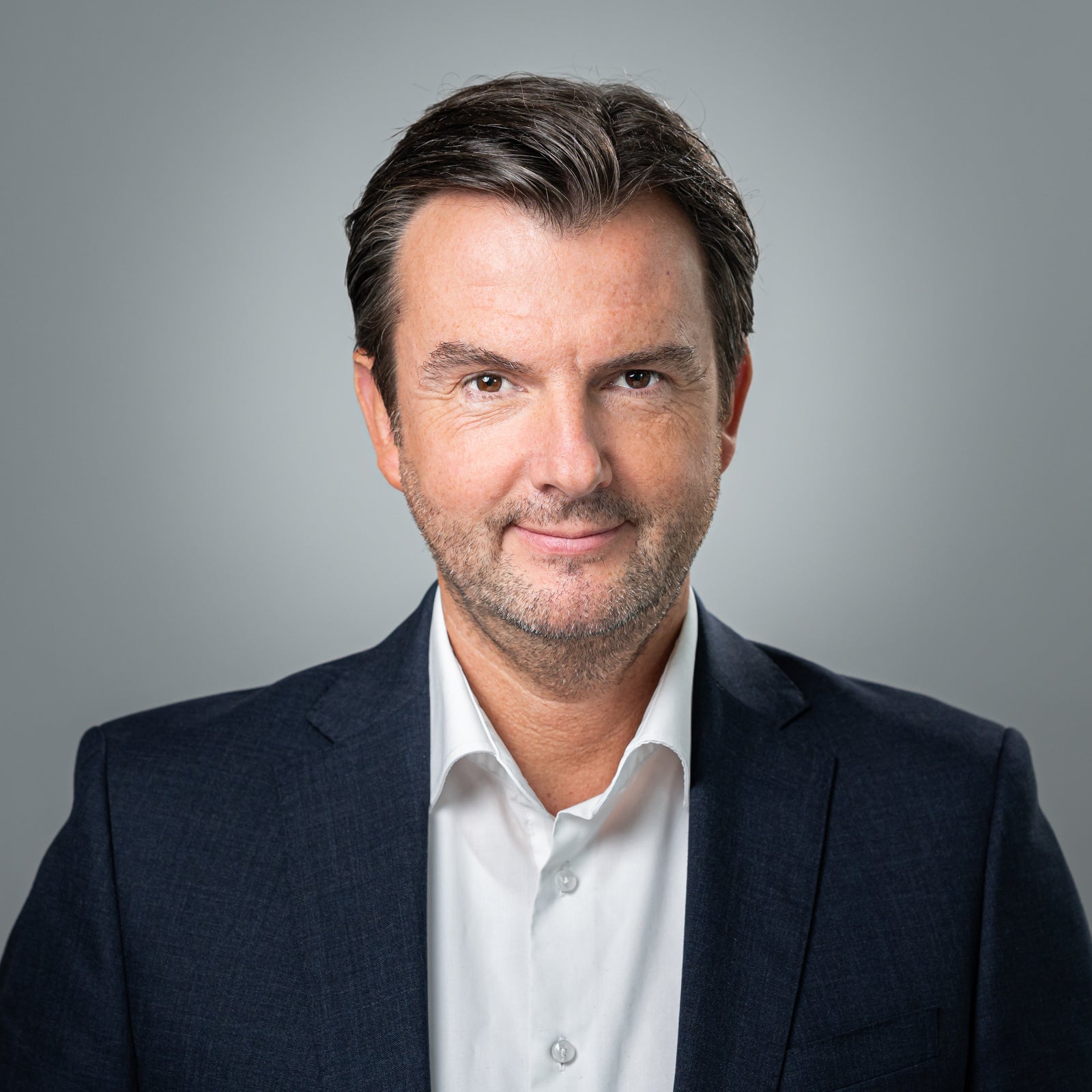 Portrait Klaus Markus. Mann mit Anzug lächelt in die Kamera, grauer Hintergrund.