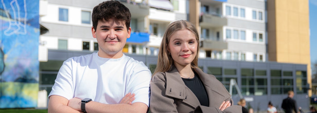 Zwei Azubis in der Ausbildung für Immobilienkaufleute stehen lächelnd und mit verschränkten Armen vor einem Vonovia Gebäude.