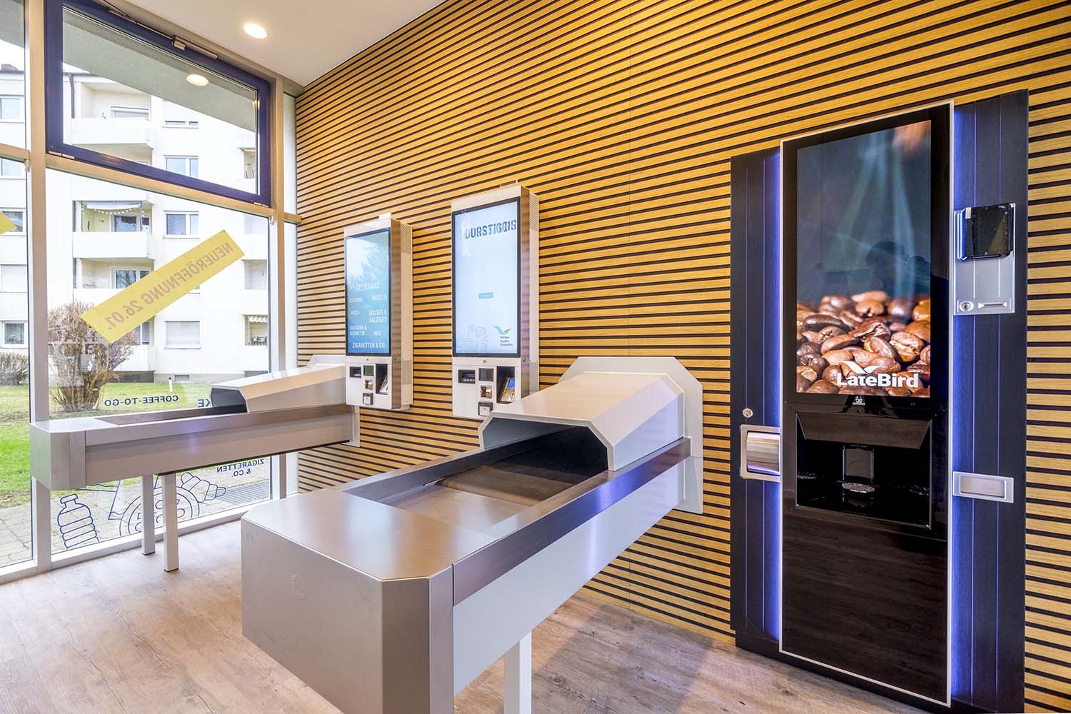 Das Bild zeigt einen modernen Raum mit Selbstbedienungsautomaten. An der Wand befinden sich zwei Bedienfelder, daneben ein Kaffeeautomat. Es gibt zwei rechteckige Tische mit Transportbändern. Der Raum hat Holzleisten an der Wand und große Fenster mit Blick auf Wohngebäude und eine Rasenfläche.