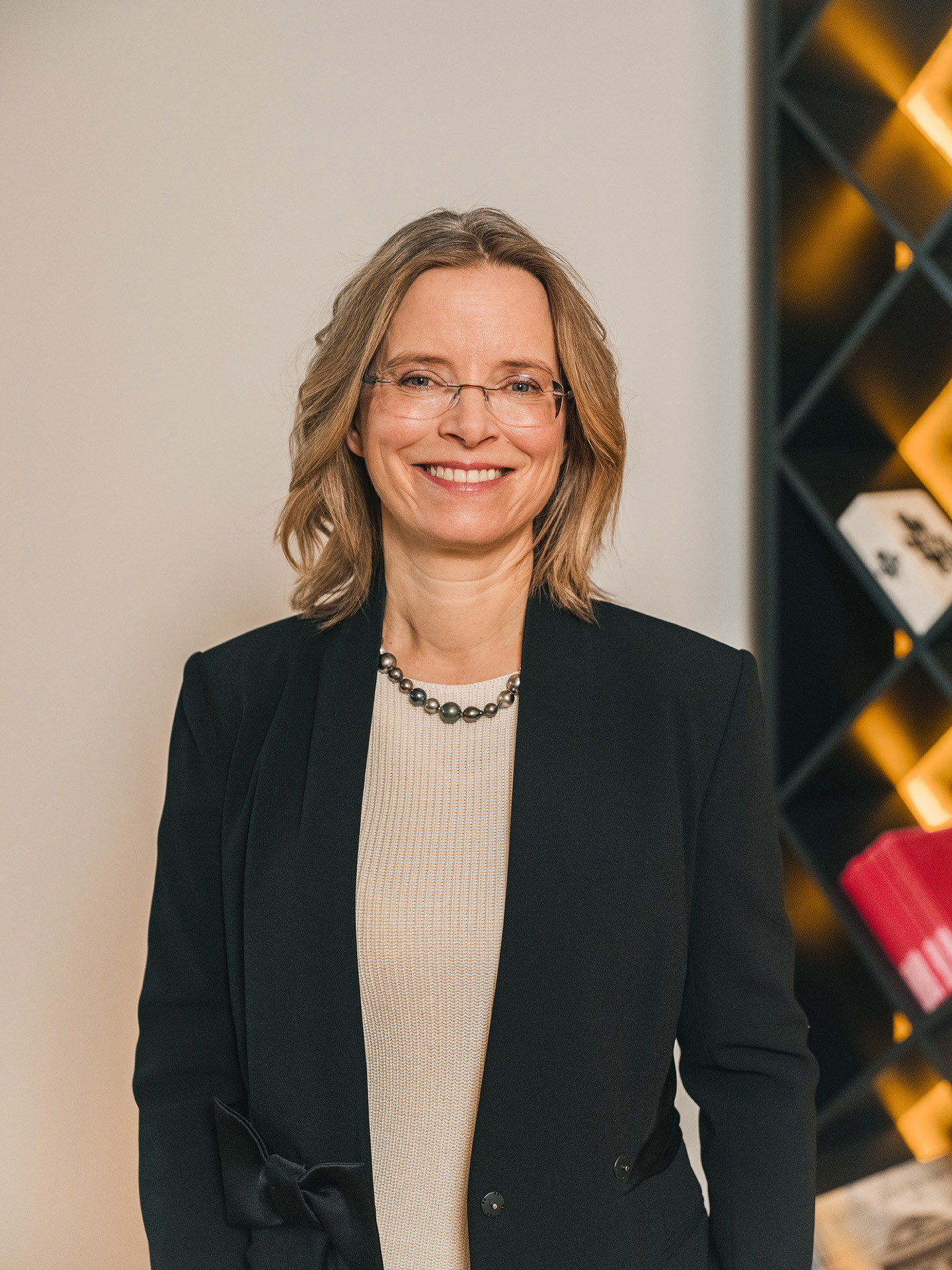 Portrait Birgit M. Bohle. Frau, in Businesskleidung lächelt und steht in einem Büro mit einem Regal. Portrait Birgit M. Bohle. Frau, in Businesskleidung lächelt und steht in einem Büro mit einem Regal.