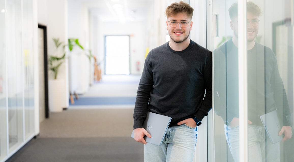 Ein Auszubildender mit einem Notebook in der Hand steht in einem Büro.