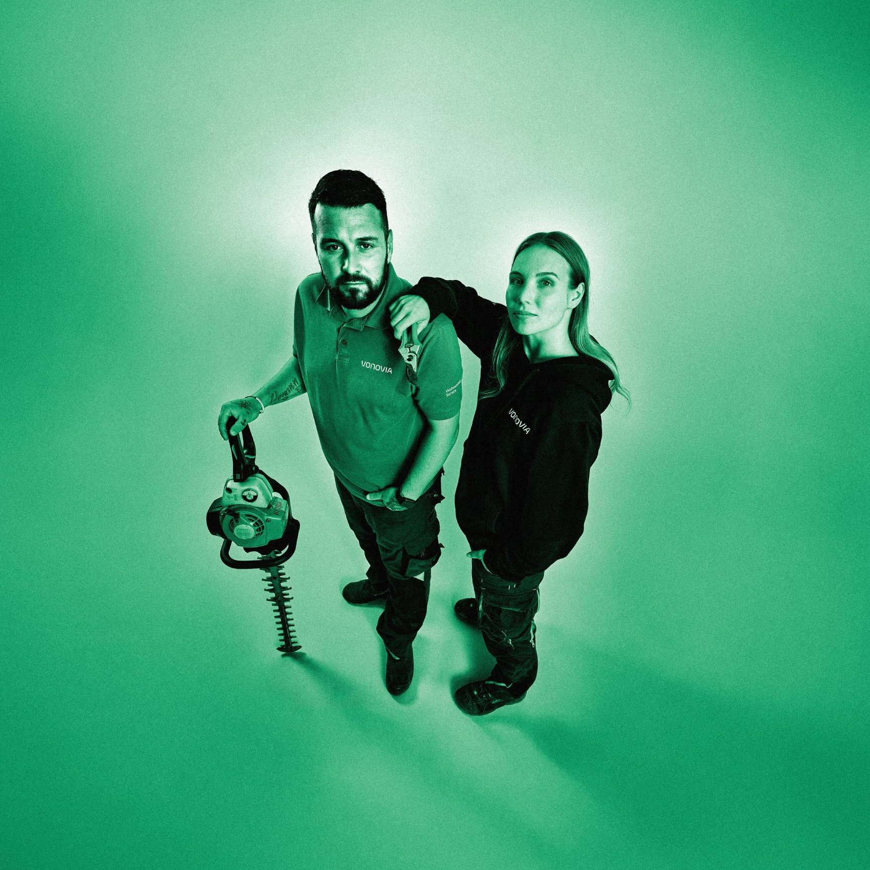 Zwei Mitarbeitenden im Gartenbau schauen nach oben in die Kamera und posieren mit einer Kettensäge. Grüner Filter über dem gesamten Bild.