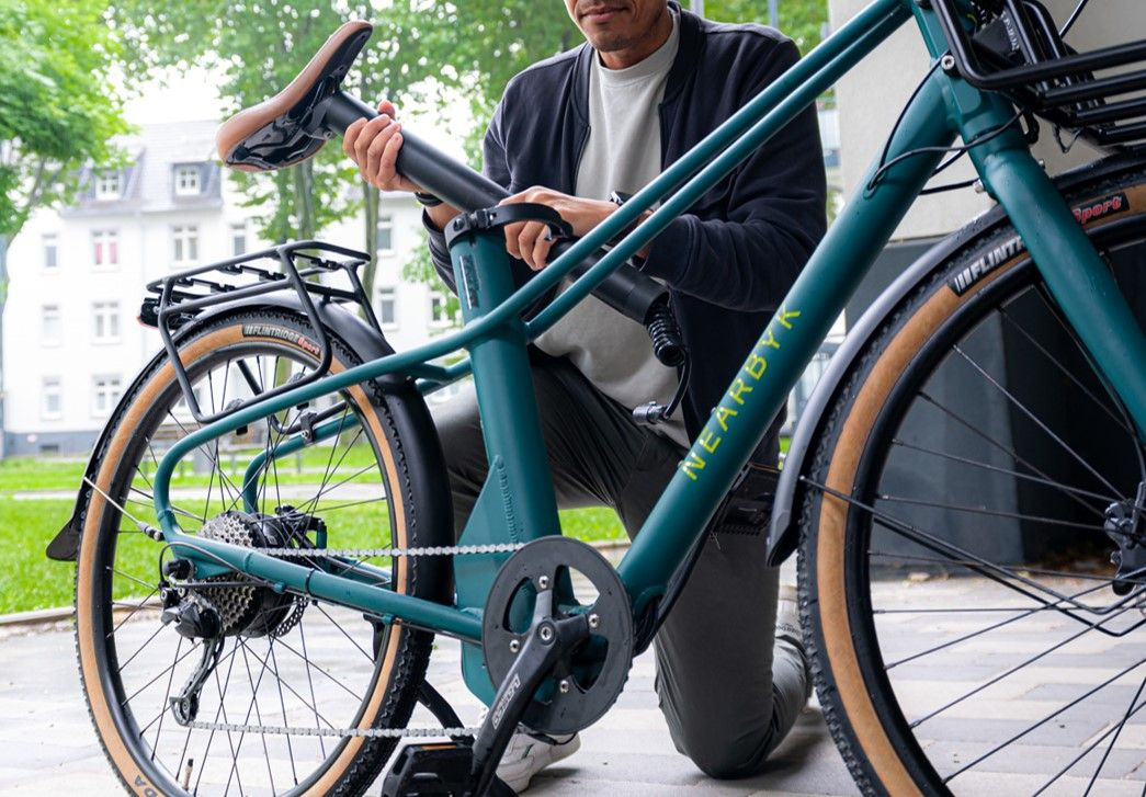 Ein Mann stellt den Sattel eines grünen Fahrrads der Marke Nearbyk mit einem Wohngebiet im Hintergrund ein. Er scheint sich auf seine Aufgabe zu konzentrieren und kniet neben dem Fahrrad auf einer gepflasterten Fläche. Ein Mann stellt den Sattel eines grünen Fahrrads der Marke Nearbyk mit einem Wohngebiet im Hintergrund ein. Er scheint sich auf seine Aufgabe zu konzentrieren und kniet neben dem Fahrrad auf einer gepflasterten Fläche.