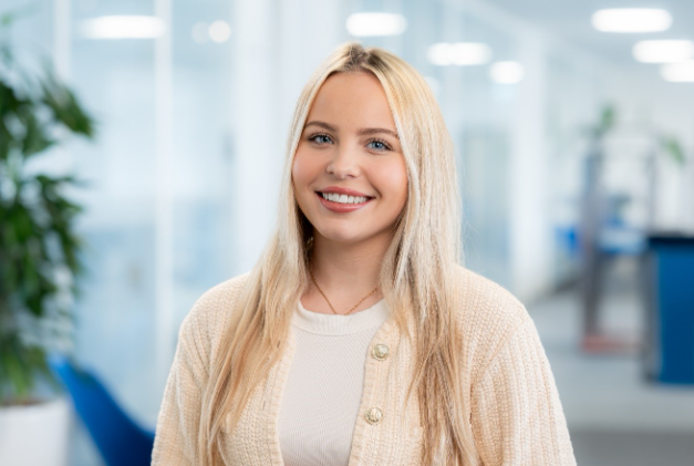 Portrait Jaclyn Schikora. Frau lächelt in einem Büro mit Pflanzen und moderner Einrichtung.
