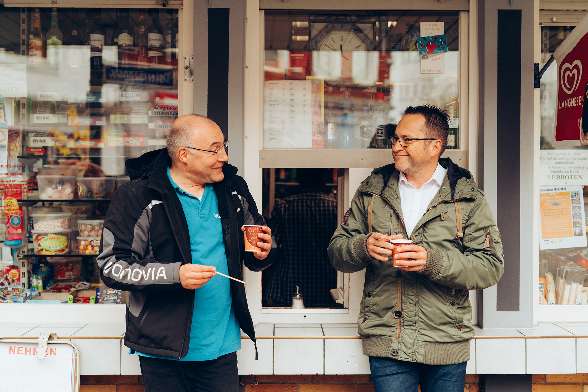 Zwei Männer unterhalten sich und trinken Kaffee vor einem Kiosk.