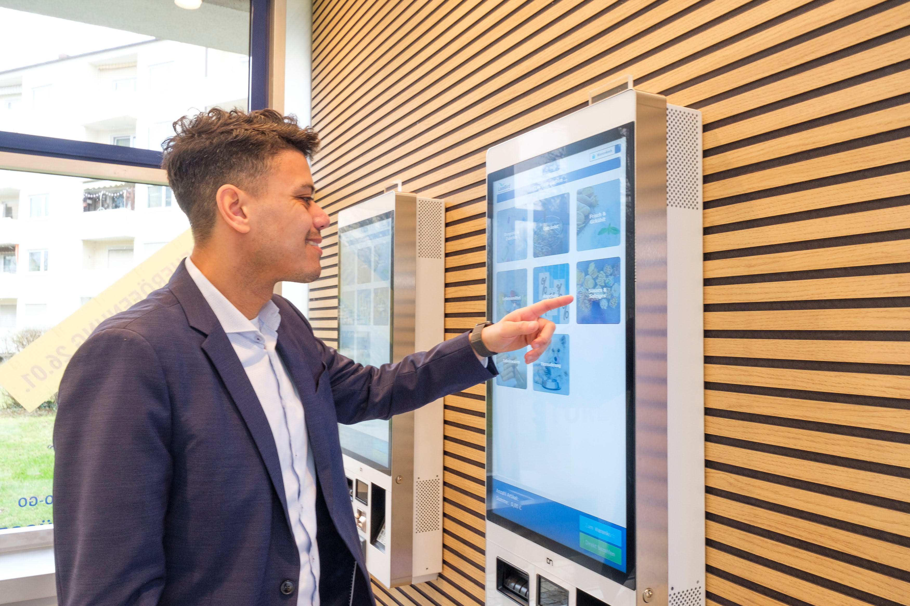 Ein Mann steht vor einem großen Touchscreen in einem modernen Raum mit Holzleisten an der Wand. Er trägt einen dunkelblauen Anzug und tippt auf dem Bildschirm, auf dem verschiedene Artikel und Optionen angezeigt werden.