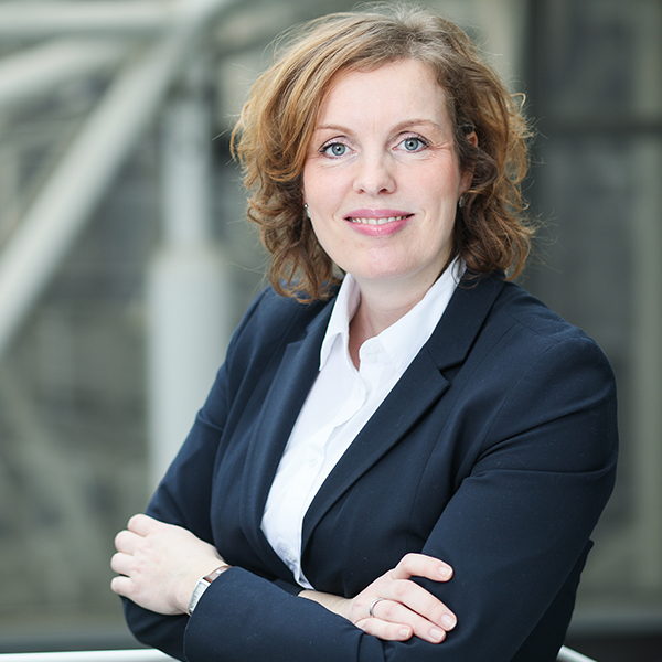 Portrait Silke Ternes. Frau steht lächelnd mit verschränkten Armen vor verschwommenem Hintergrund.