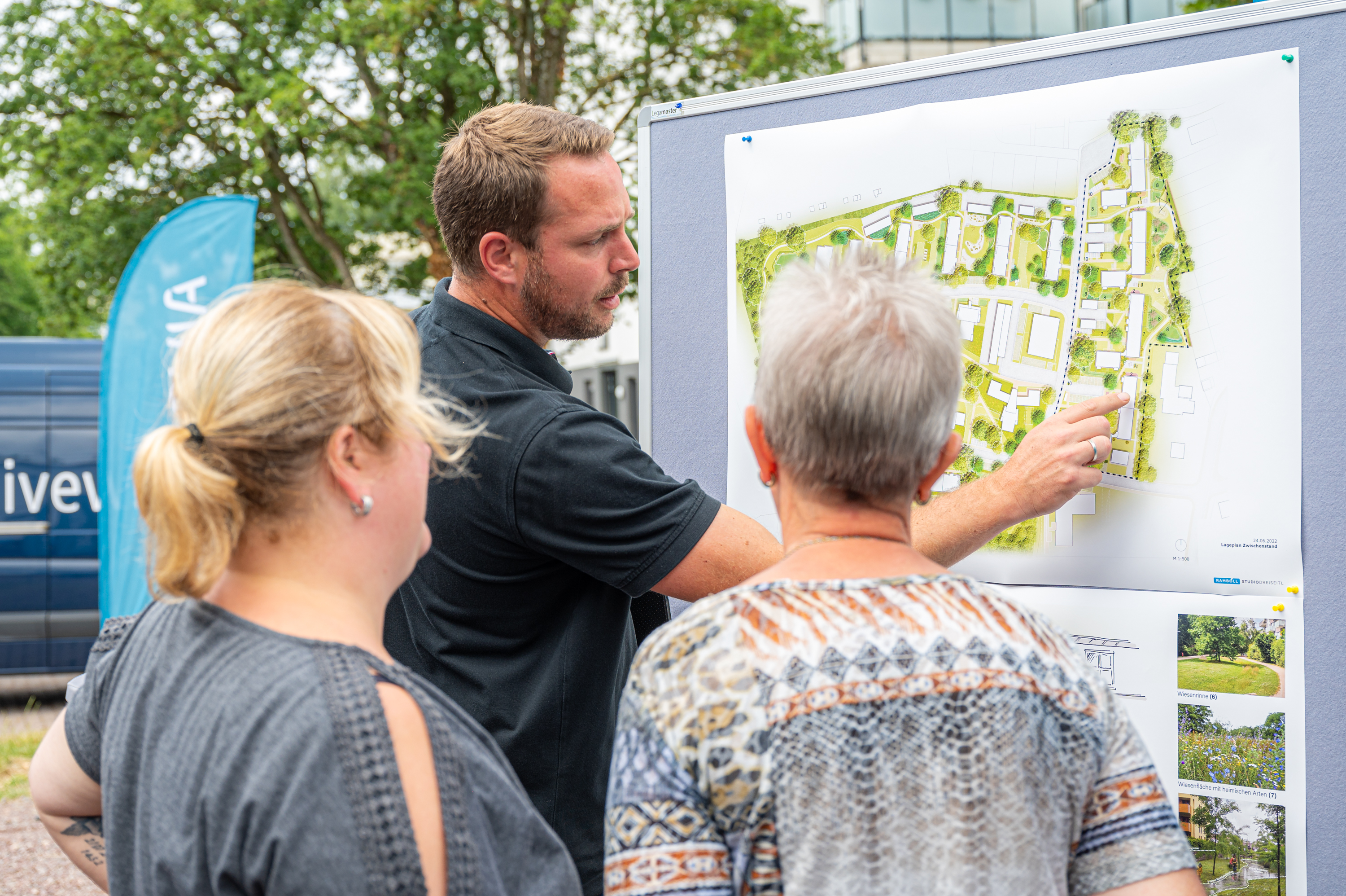 Mann zeigt zwei Frauen einen Bauplan auf einem Informationsstand.