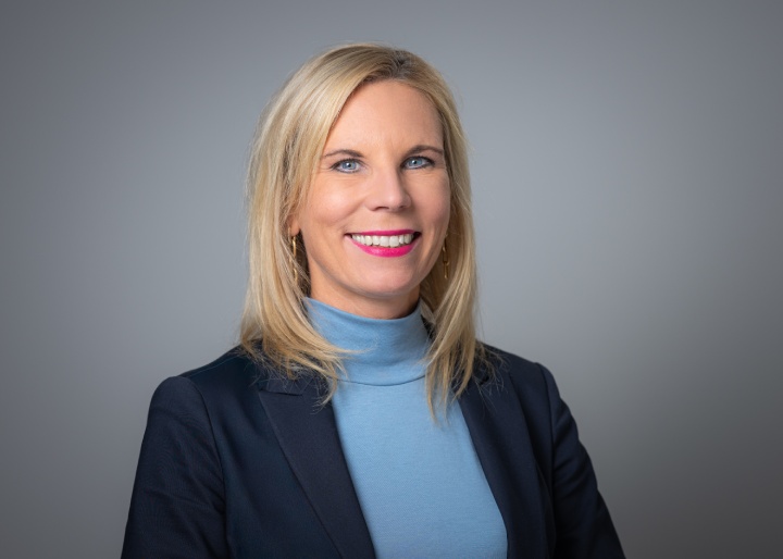 Ein Foto von Christina Jordan. Frau mit blonden Haaren mit einem hellblauen Rollkragenpullover und einen dunkelblauen Blazer vor einem grauen Hintergrund.