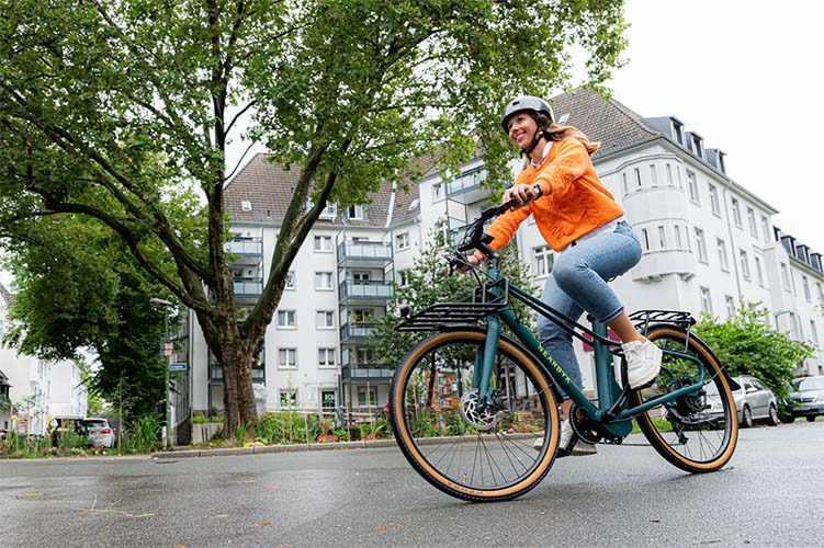 Eine Frau in einer orangefarbenen Jacke und Jeans fährt lächelnd ein grünes NEARBYK Fahrrad durch eine städtische Wohngegend. Sie trägt einen Fahrradhelm, und im Hintergrund sind Wohnhäuser sowie ein großer Baum zu sehen. Eine Frau in einer orangefarbenen Jacke und Jeans fährt lächelnd ein grünes NEARBYK Fahrrad durch eine städtische Wohngegend. Sie trägt einen Fahrradhelm, und im Hintergrund sind Wohnhäuser sowie ein großer Baum zu sehen.