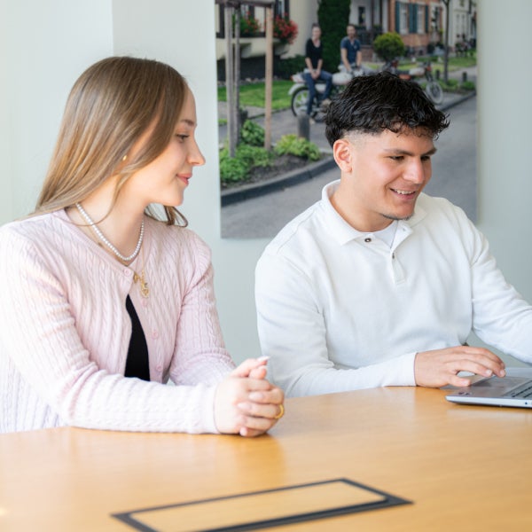 Zwei Auszubildende im Dualen Studium sitzen an einem Schreibtisch und schauen lächelnd auf ein Notebook.