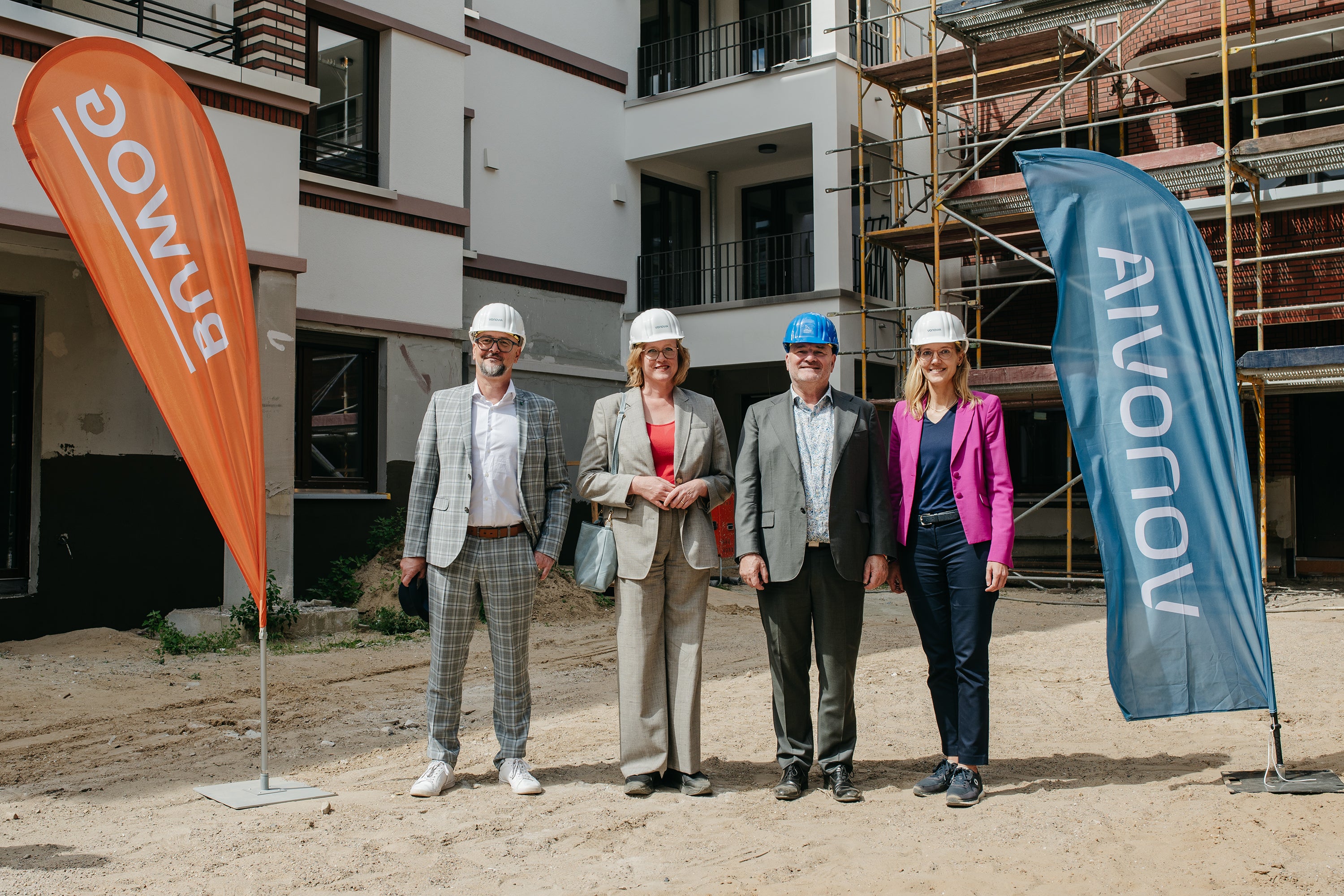 Vier Personen mit Helmen stehen auf einer Baustelle zwischen zwei Bannern. Ein Banner ist orange mit dem Schriftzug BUWOG, der andere Banner ist blau mit dem Schriftzug VONOVIA.