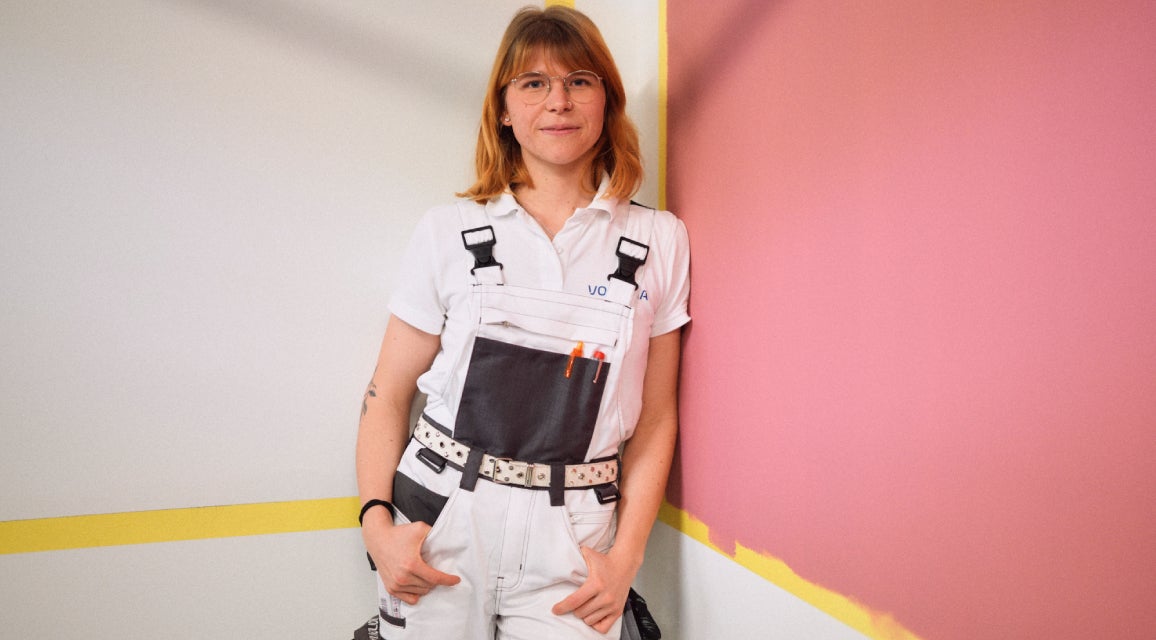 Eine Auszubildende zur Malerin & Lackiererin steht vor einer halb bemalten Wand.