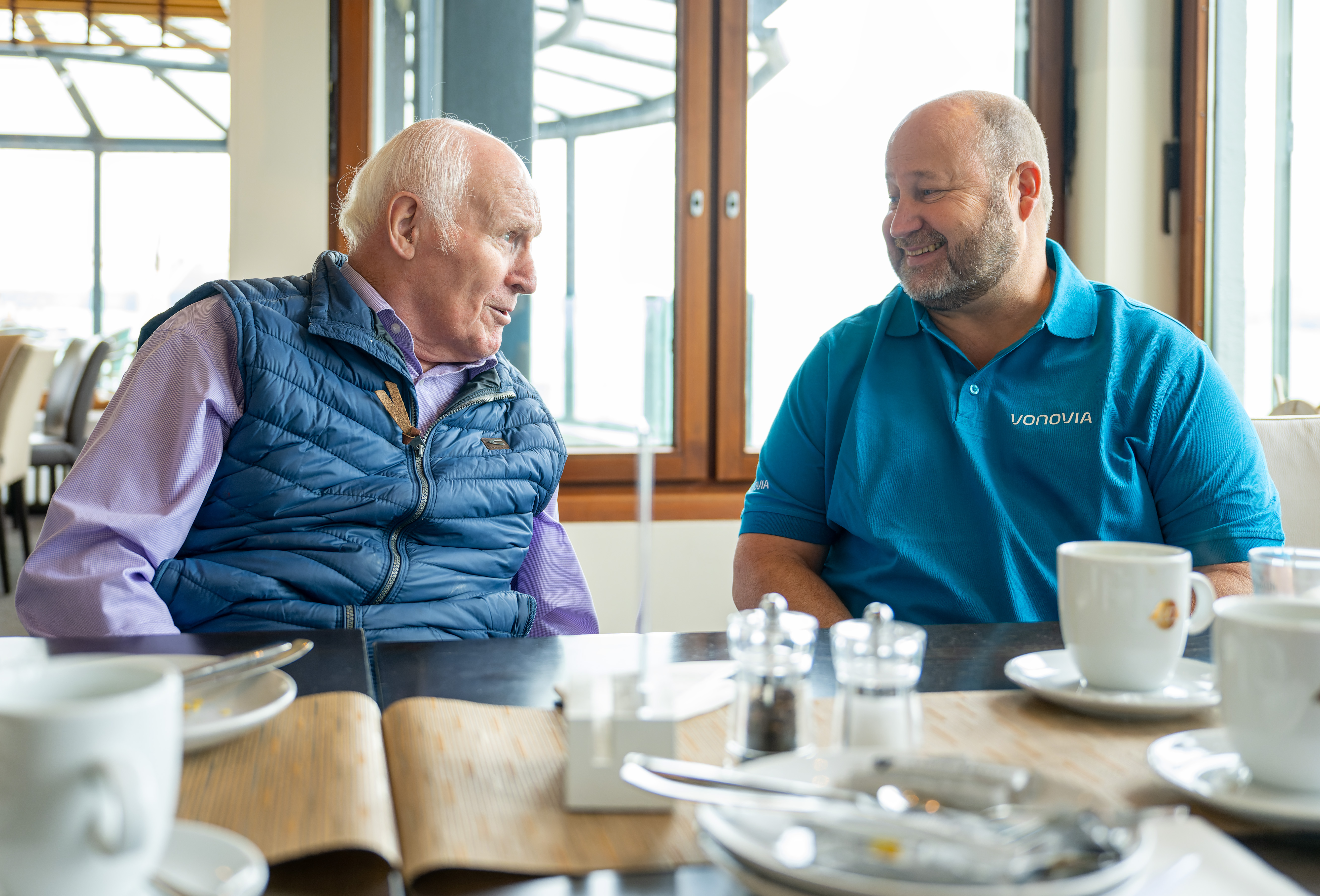 Zwei Männer sprechen in einem Café; einer trägt 