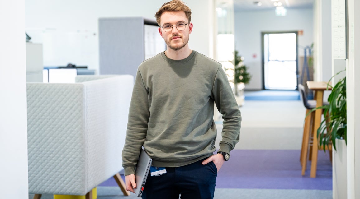 Ein Auszubildender mit einem Notebook in der Hand steht in einem Büro.