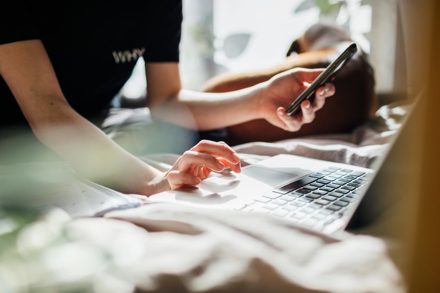 Eine Person arbeitet an einem Laptop mit einer Hand und hält in der anderen ein Smartphone. Der Hinter- und Vordergrund ist unscharf.