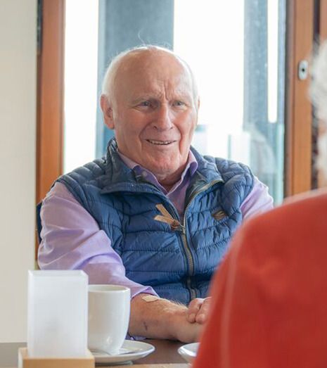 Älterer Mann sitzt und unterhält sich mit jemanden in einer Café-Umgebung.